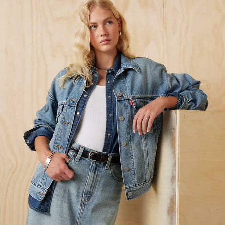 A person with long blonde hair leans against a wooden wall, wearing a 90s silhouette. Their Levis 90s Trucker Jacket drapes over a white top and matching jeans, accessorized with a black belt and silver bracelet.
