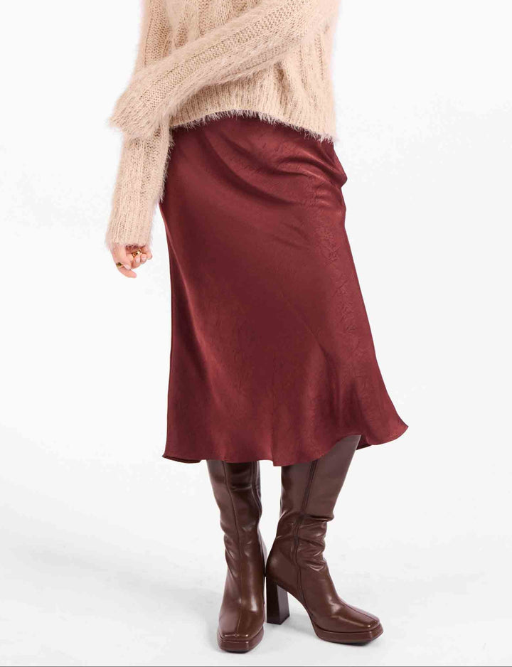 A woman modeling a fuzzy light beige knit sweater and a dark burgundy midi skirt, paired with brown knee-high boots, against a plain white studio background.
