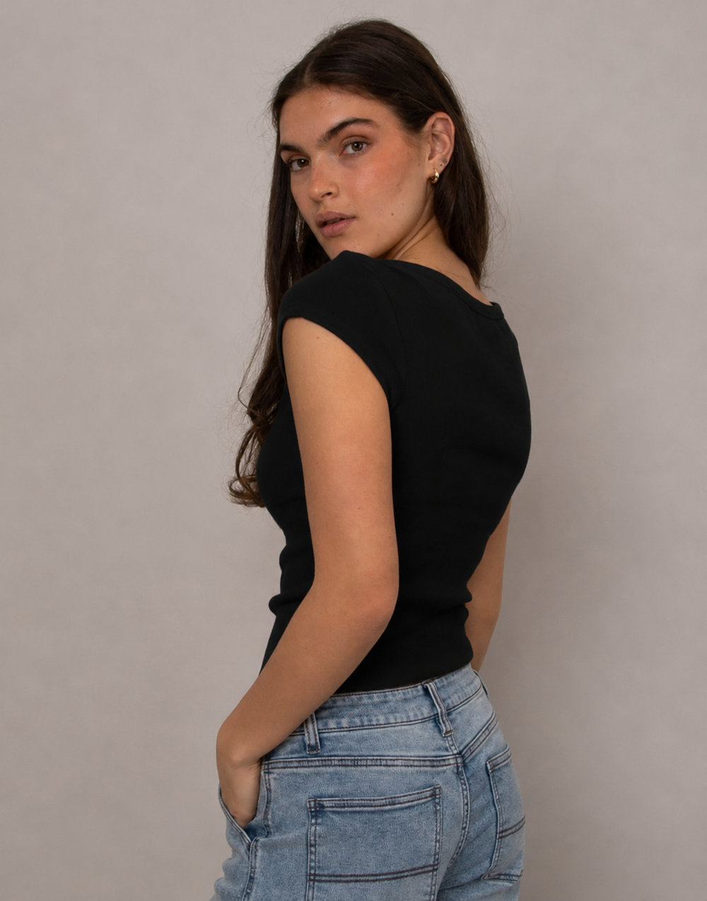 A woman with long dark hair is shown from the back wearing a black short-sleeve top and light blue jeans, standing against a plain light gray studio background.