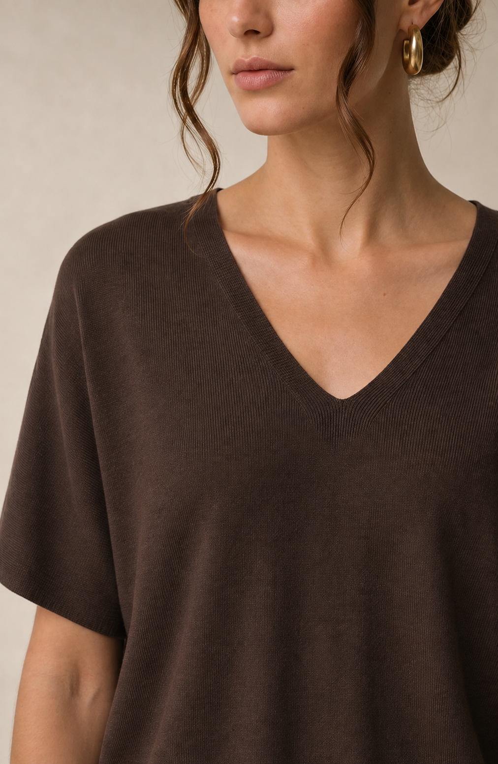 A close-up of a woman wearing a dark brown short-sleeve V-neck knit top against a neutral beige background.