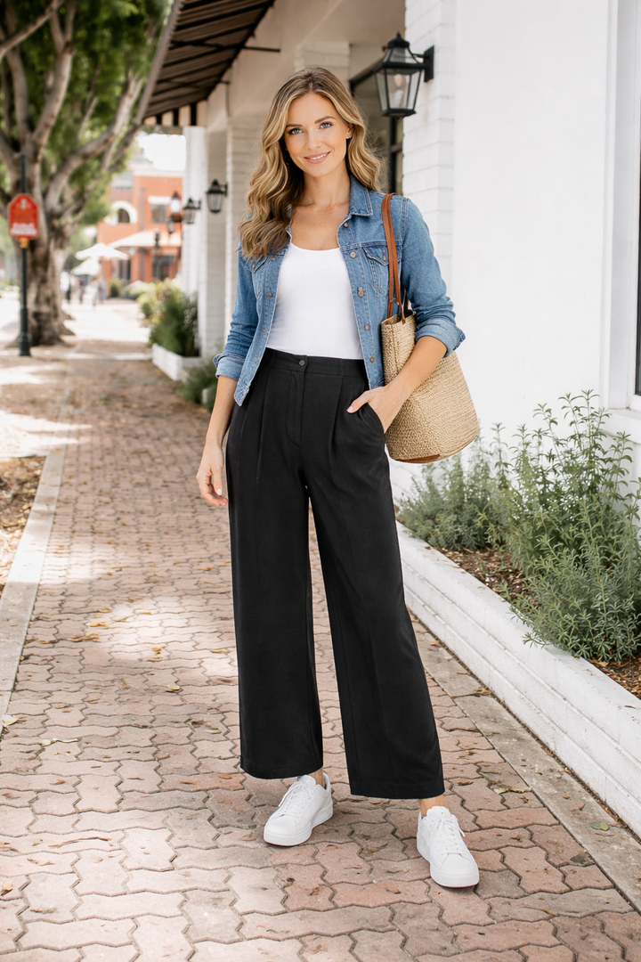 Woman walking on a sidewalk wearing a blue denim jacket, white top, black pants, and white sneakers.