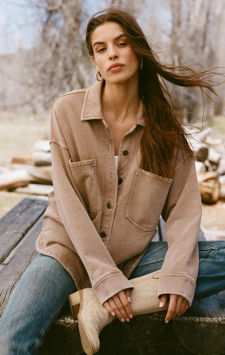 Model wearing an oversized button-up shirt in a warm, neutral tan color with two large front pockets and a casual, relaxed fit, paired with light blue denim jeans.