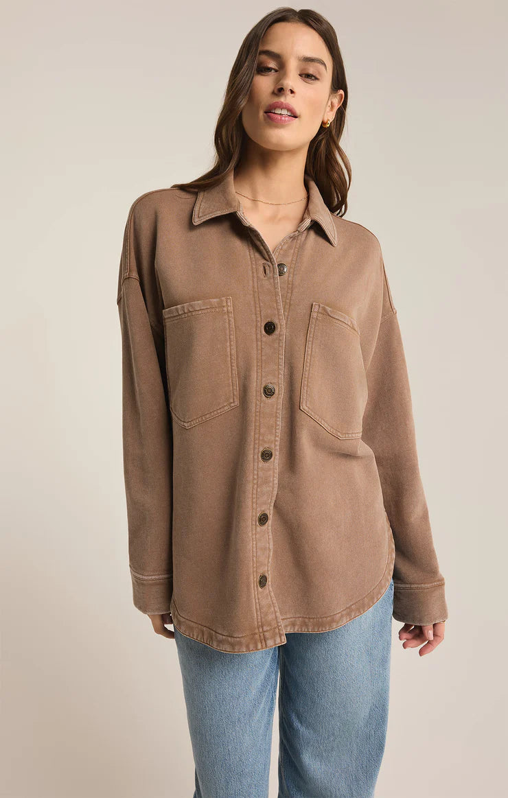 Model wearing an oversized button-up shirt in a warm, neutral tan color with two large front pockets and a casual, relaxed fit, paired with light blue denim jeans.