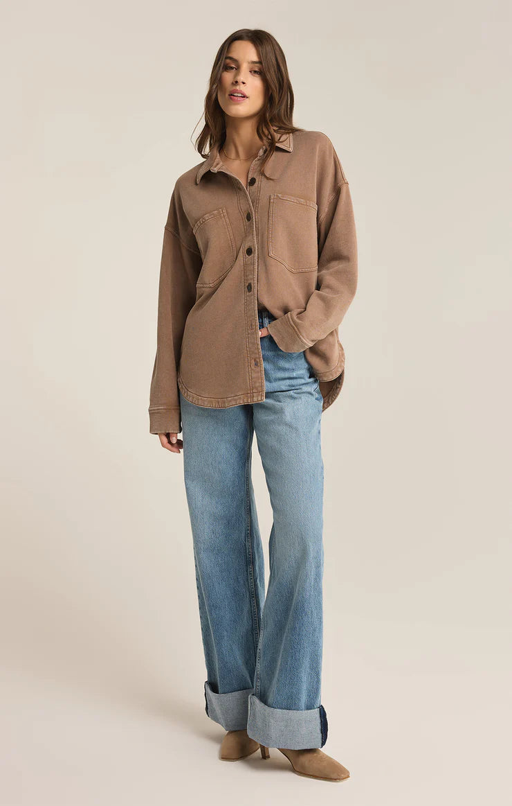 Model wearing an oversized button-up shirt in a warm, neutral tan color with two large front pockets and a casual, relaxed fit, paired with light blue denim jeans.