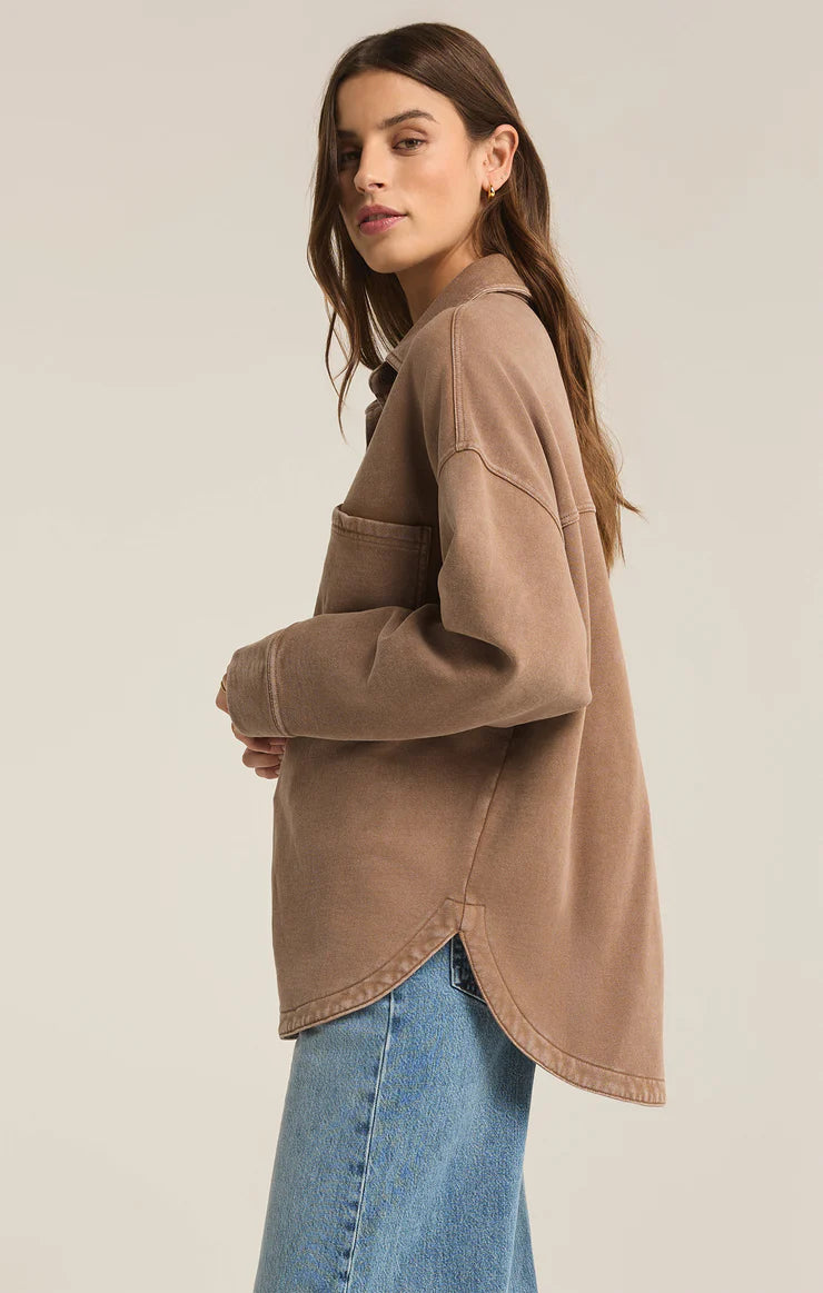 Model wearing an oversized button-up shirt in a warm, neutral tan color with two large front pockets and a casual, relaxed fit, paired with light blue denim jeans.