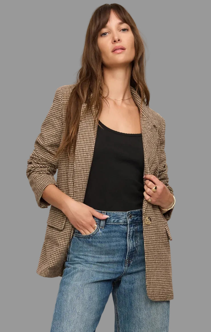 A woman wearing a brown and beige houndstooth blazer over a black tank top and blue jeans, standing against a plain light beige studio background.